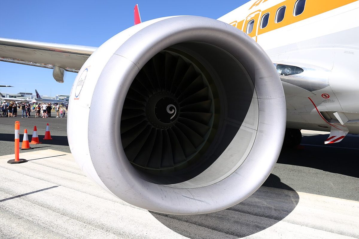 Front view of a jet engine under the wing of a parked airliner, a smooth white intake lip around a dark fan with a spiral mark on its spinner.