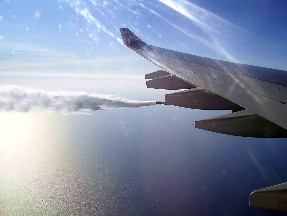 View from a cabin window of an airliner wing over the sea, a white plume streaming back from a nozzle at the wing's trailing edge.