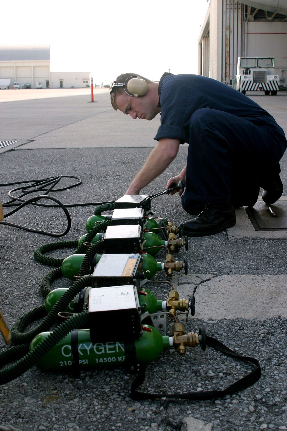 A row of small green oxygen bottles with valves and carrying straps on a ramp, a man kneeling behind them.
