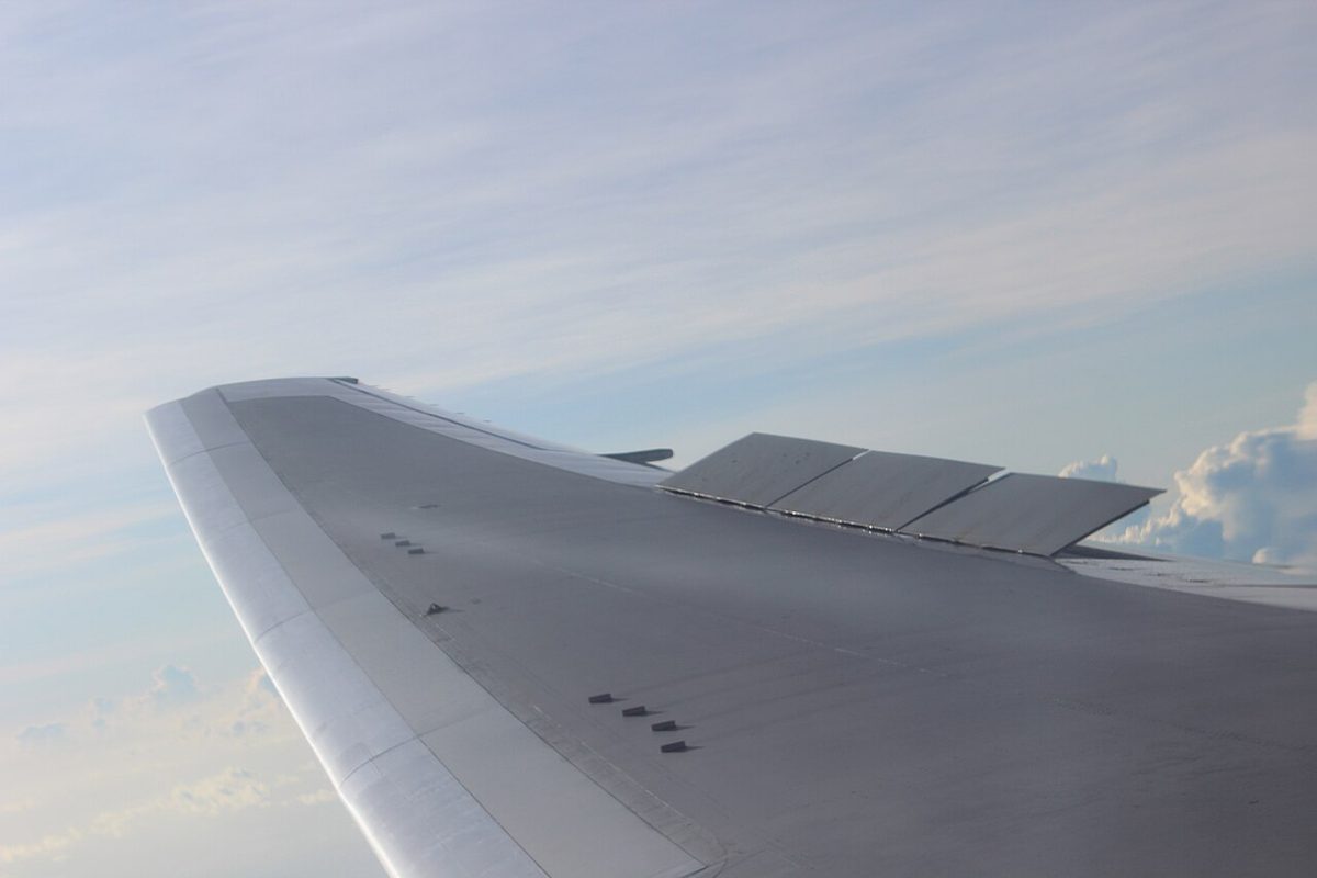 View from a cabin window of an airliner's right wing in flight, with several spoiler panels partly raised from the upper surface.