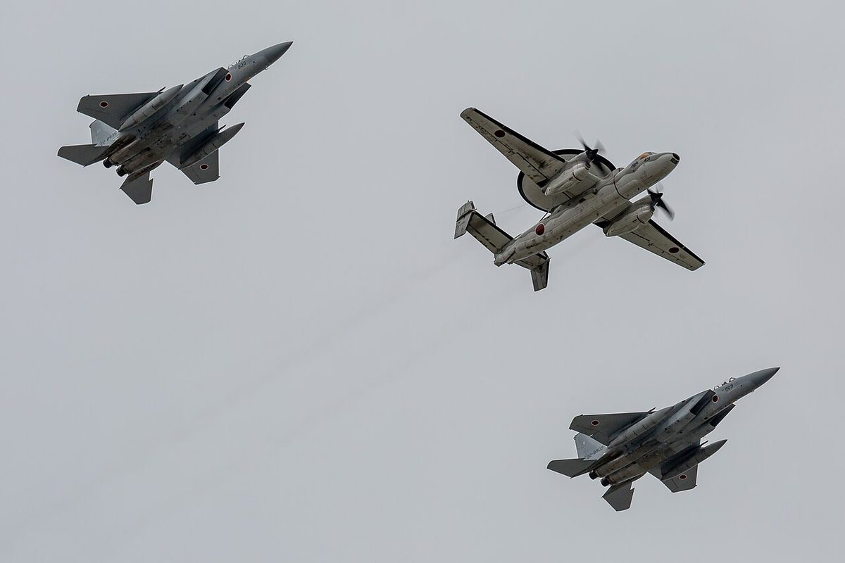 Two F-15 fighters flying in formation on either side of a twin-propeller E-2 radar aircraft, seen from below against a grey sky.