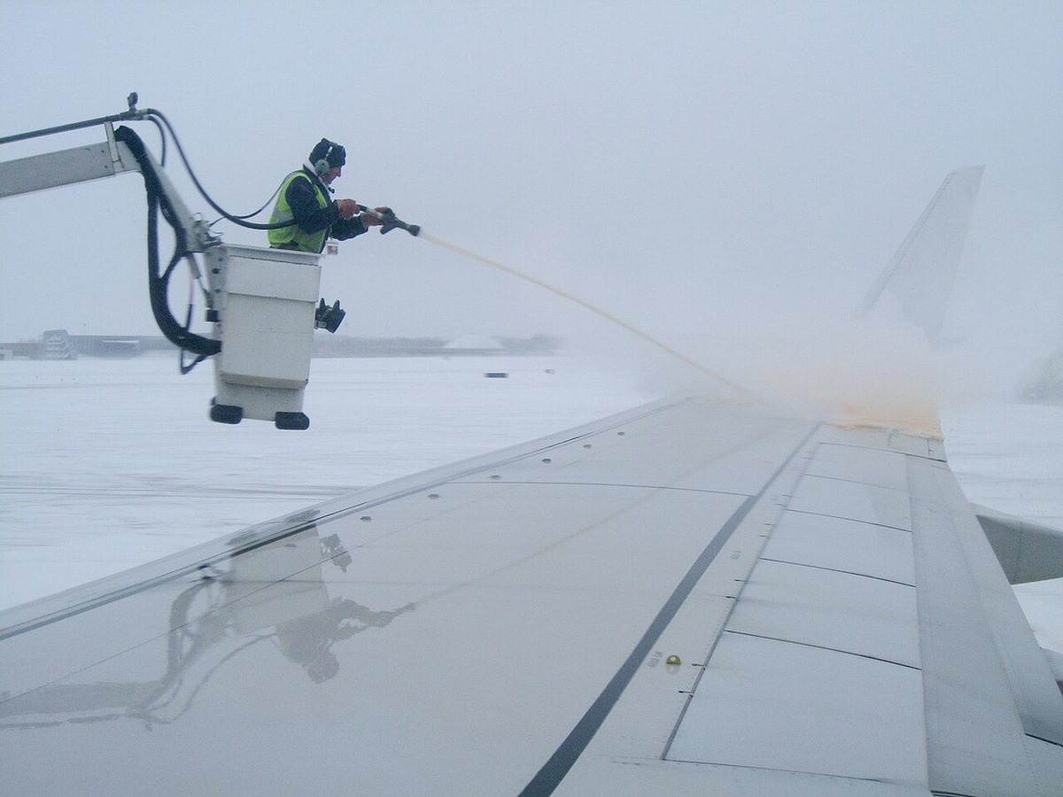 A worker in the raised basket of a de-icing truck sprays fluid across an airliner's snowy wing, seen from a cabin window.