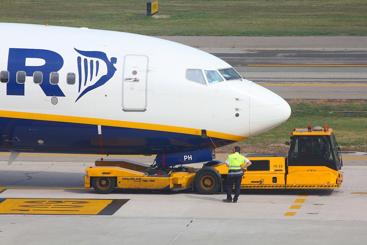 The nose of a Boeing 737 held in a low yellow towbarless tractor on an apron, a ground worker in a high-visibility vest standing beside it.