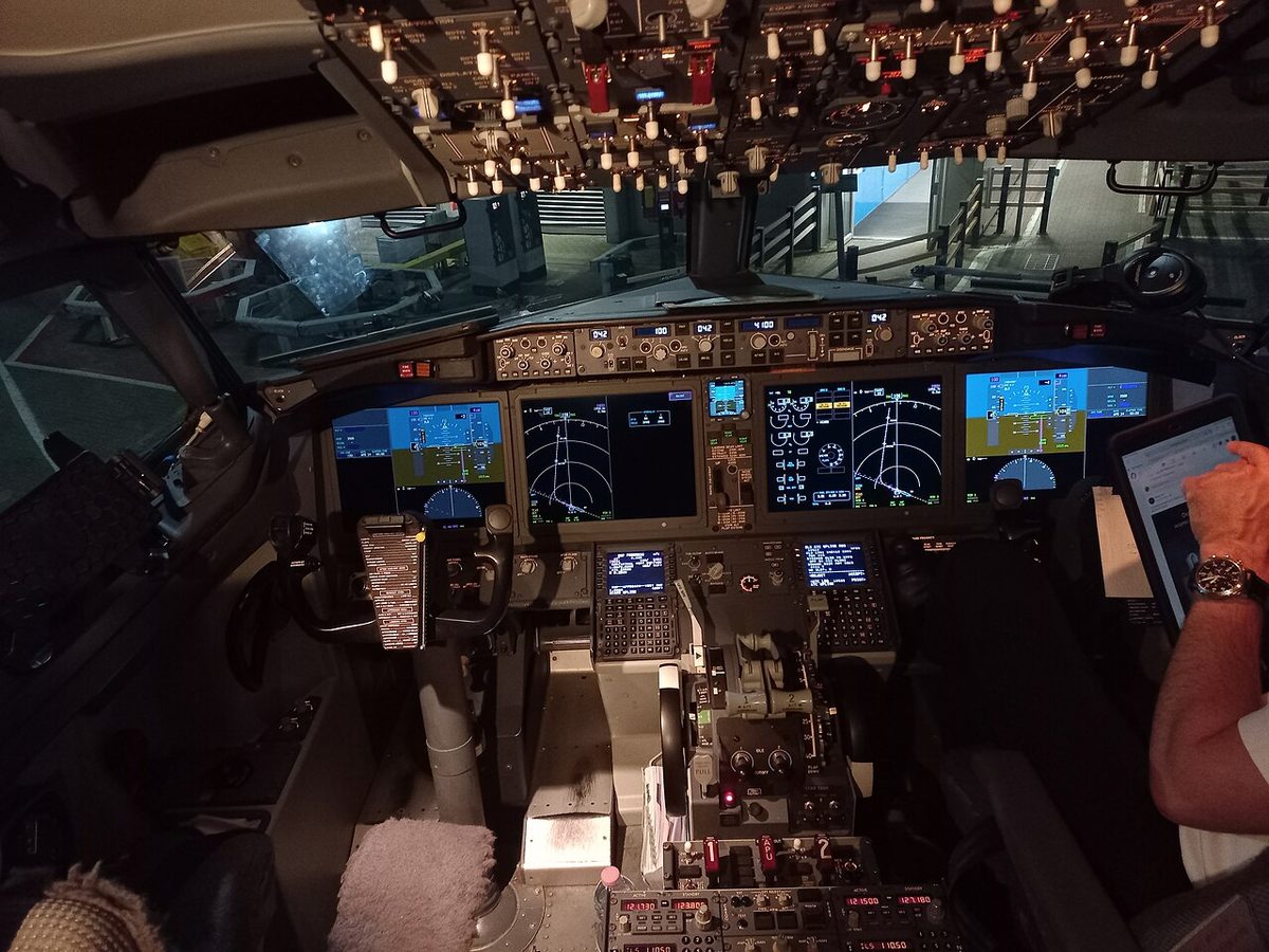 A dim Boeing 737 MAX flight deck at night seen from behind the seats: four large lit screens, the control columns, a lit overhead panel and, at right, a pilot's hand on a tablet.