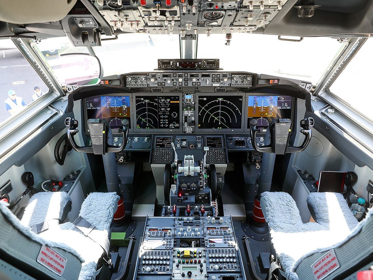 Airliner flight deck seen from behind two sheepskin-covered seats, four large display screens across the main panel.