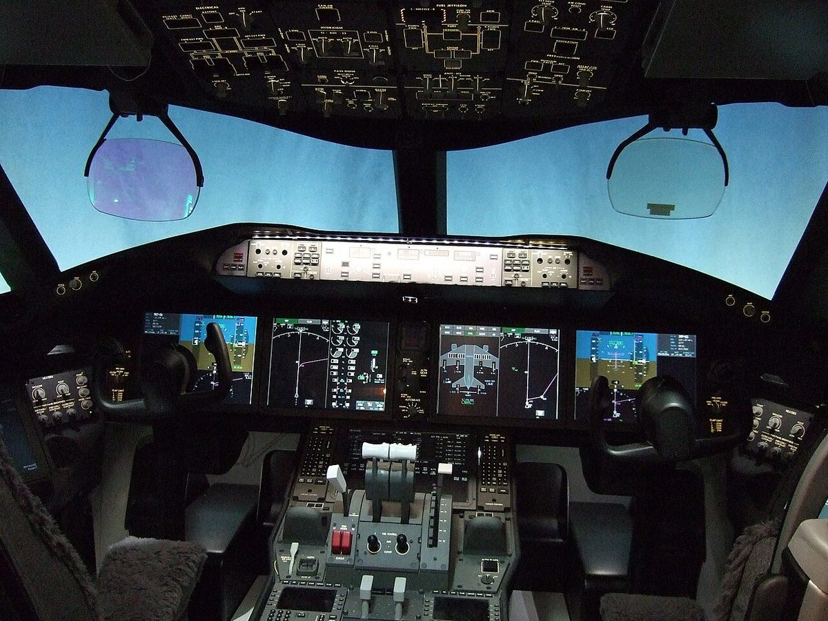 A Boeing 787 flight deck: two head-up display combiners lowered in front of the pilot seats, large display screens across the main panel and the overhead panel above.