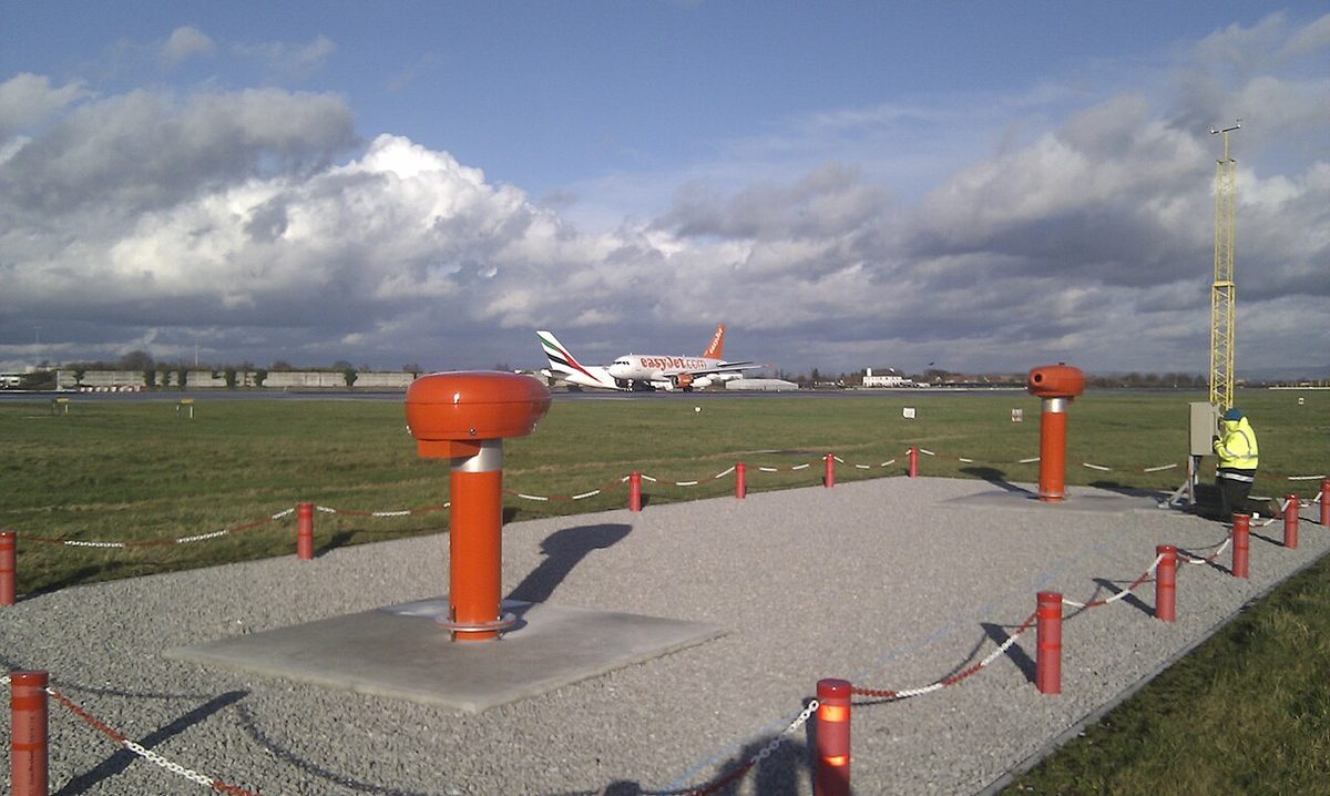 Orange instrument housings on posts, set in gravel beside a grassy airfield, with a parked airliner and a cloudy sky beyond.