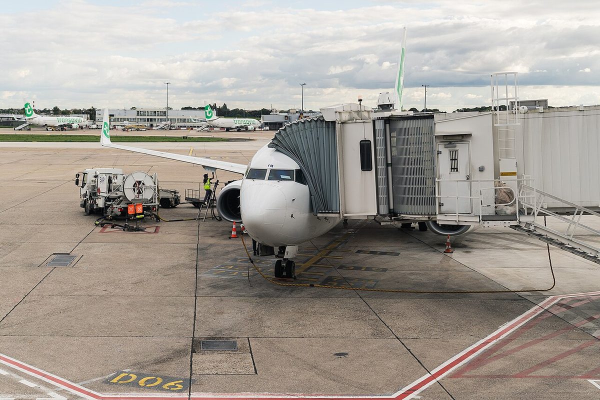 A Transavia Boeing 737-800 at a jet bridge, with a fuel vehicle parked beside it, hoses connected, and a worker in a high-visibility vest on a stand next to the engine.