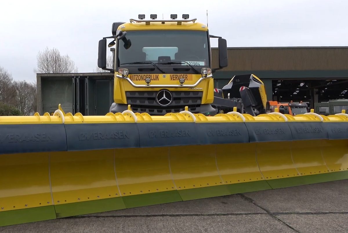 A yellow truck fitted with a very wide yellow snow plough blade, parked on concrete in front of a hangar.