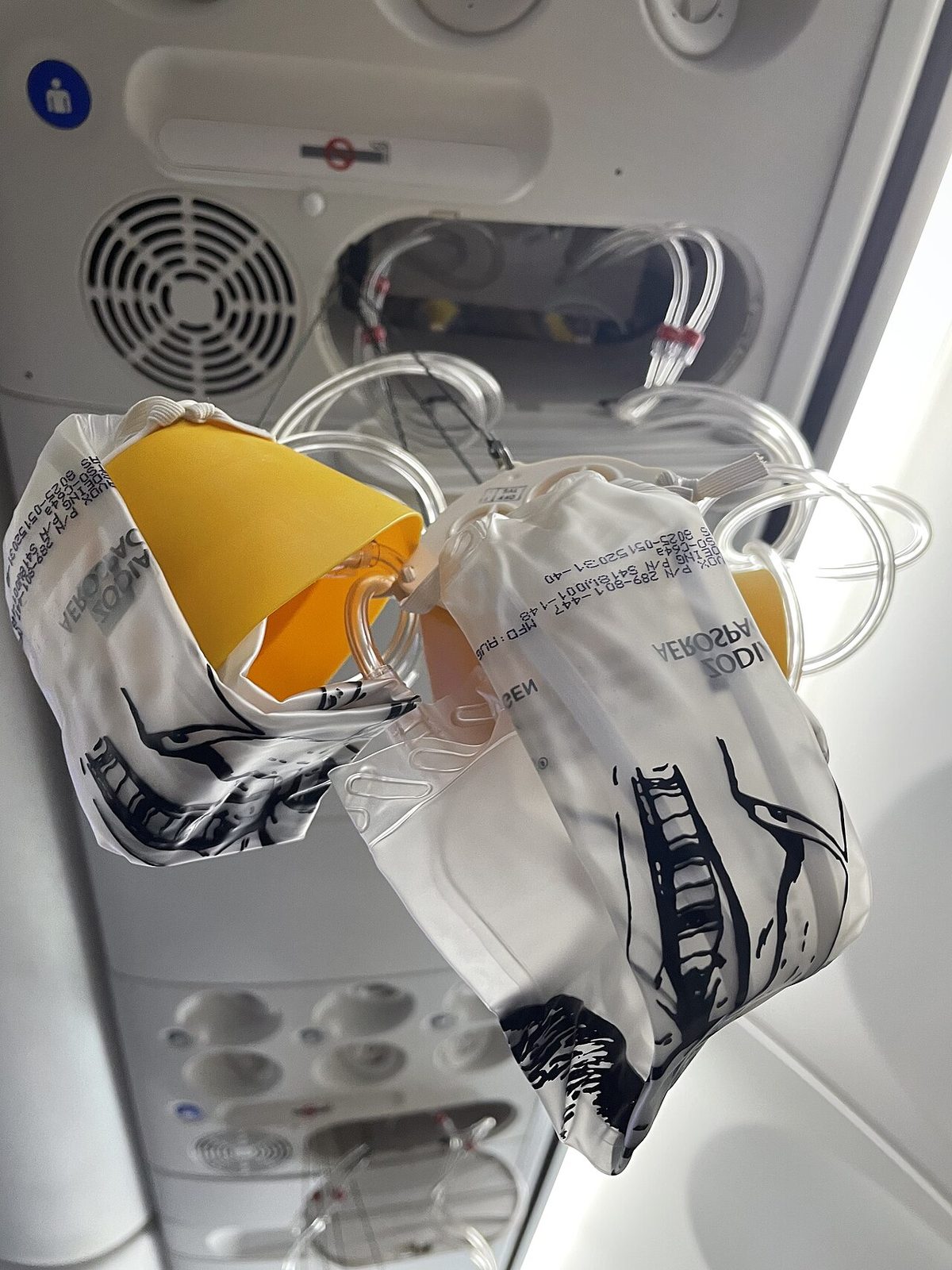 Clear plastic oxygen masks with yellow cups and reservoir bags hanging on tubes from an open overhead panel in an airliner cabin.
