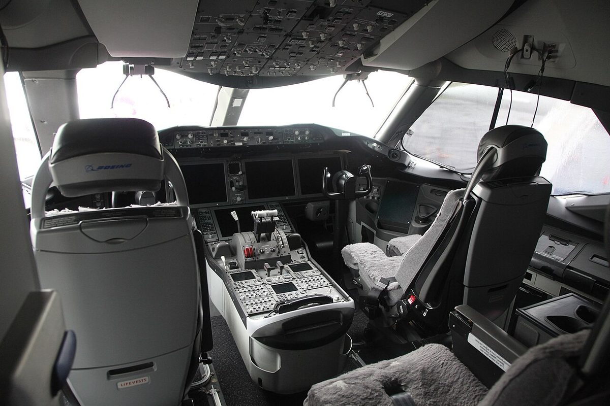 A Boeing 787 flight deck seen from behind the two seats: control wheels, the centre pedestal between the seats and the overhead panel above, the displays switched off.