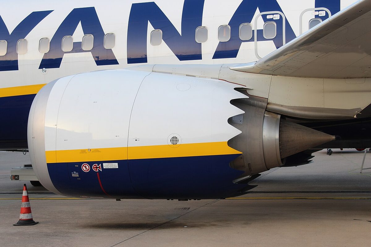 Side view of a large white and dark blue engine nacelle with a saw-tooth rear edge, mounted close under the wing of a parked airliner.