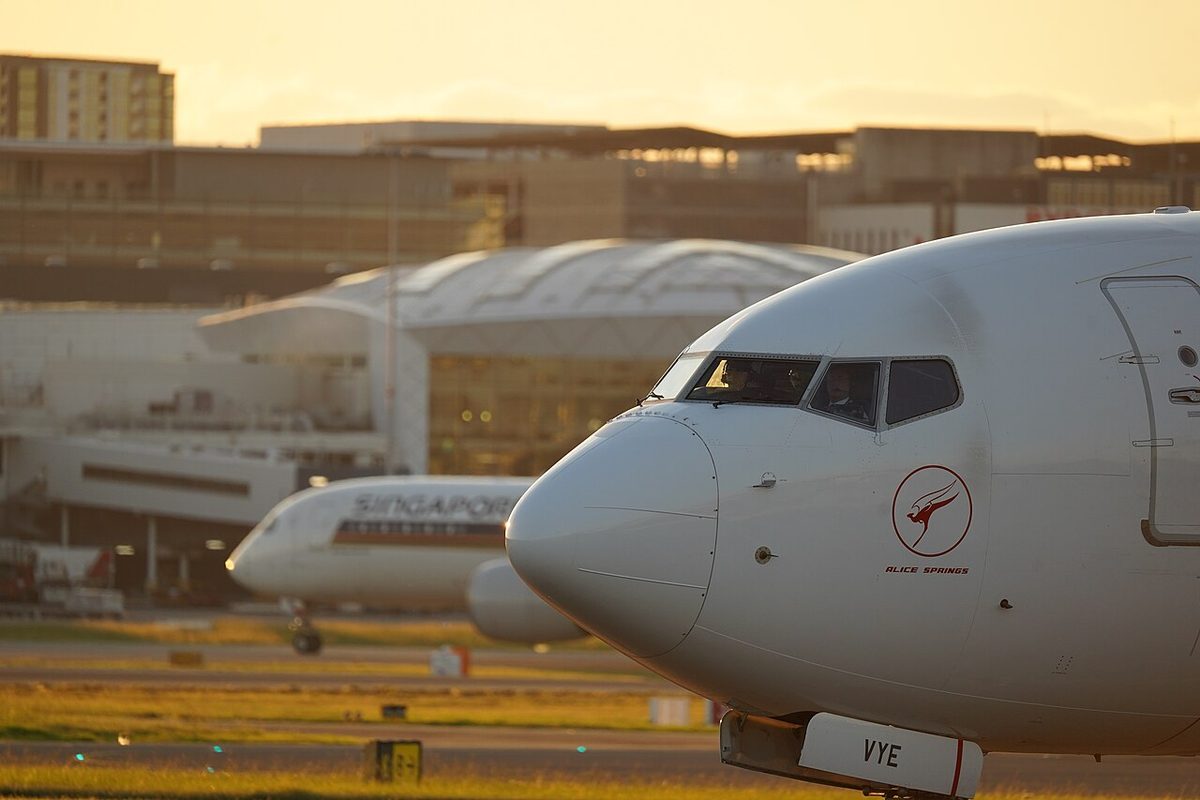 The nose of a white airliner at sunset, seen from the left side, with its three flight deck windows and the pilots faintly visible inside.