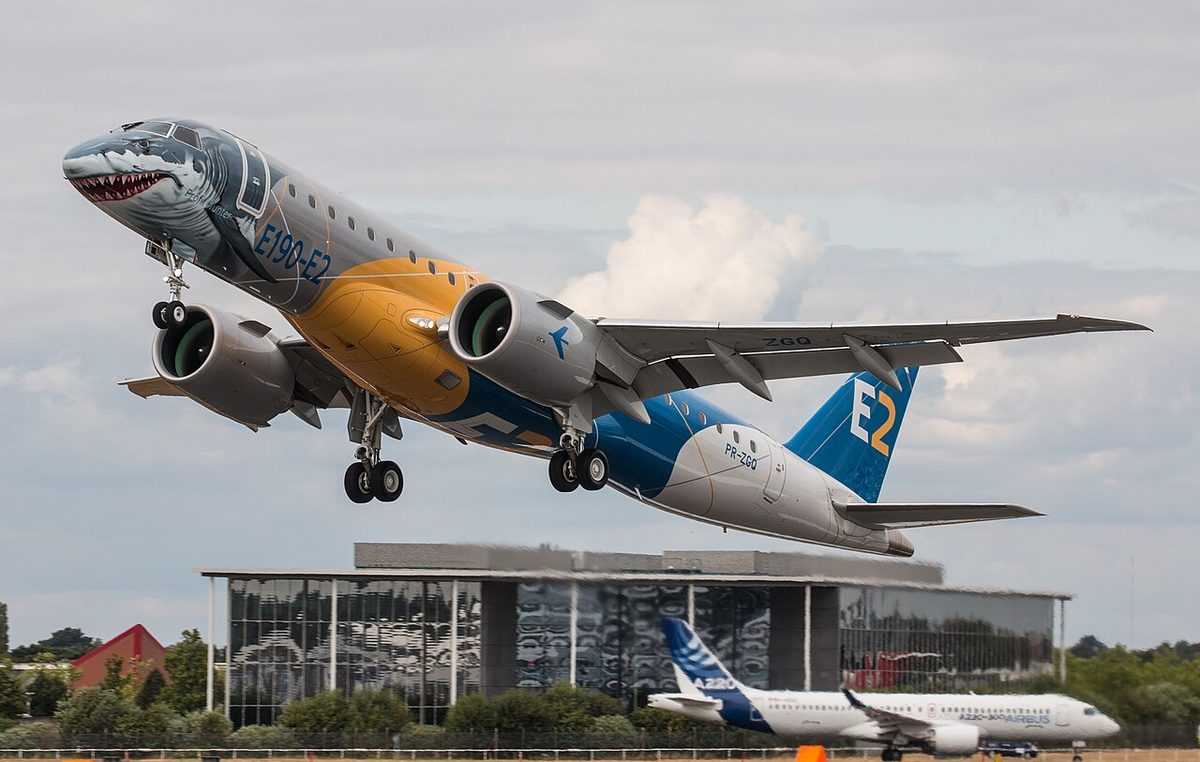 A twin-engined jet with a shark's face painted on its nose climbing steeply after take-off, gear down, its long, slender right wing stretched out.
