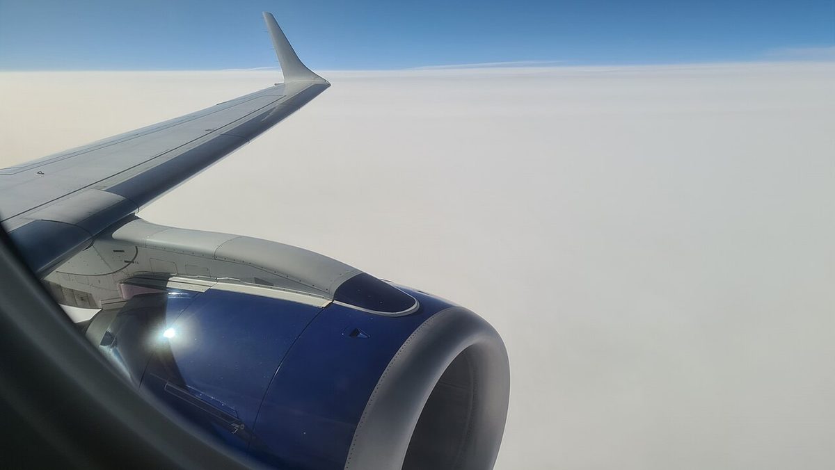 A cabin window view of an airliner wing with an upturned tip and a dark blue engine nacelle, above a layer of cloud.