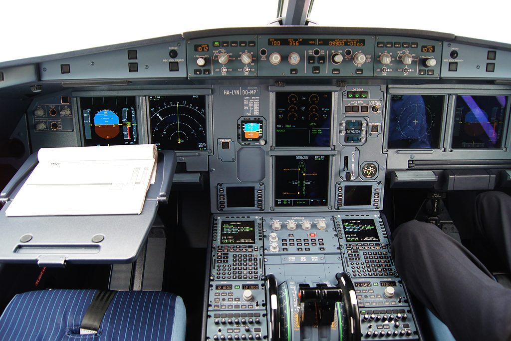 Airbus flight deck seen from behind the seats: two screens stacked in the center panel, the lower one showing a fuselage outline with small amber squares, the captain's flight displays on the left and the pedestal below.