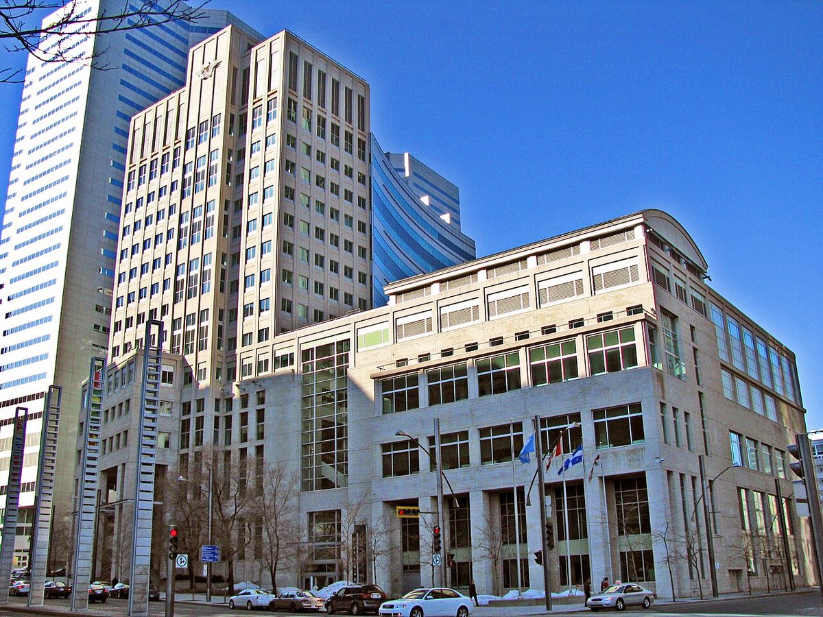 A pale stone office building with flags in front of it and a tall tower behind, under a deep blue sky.