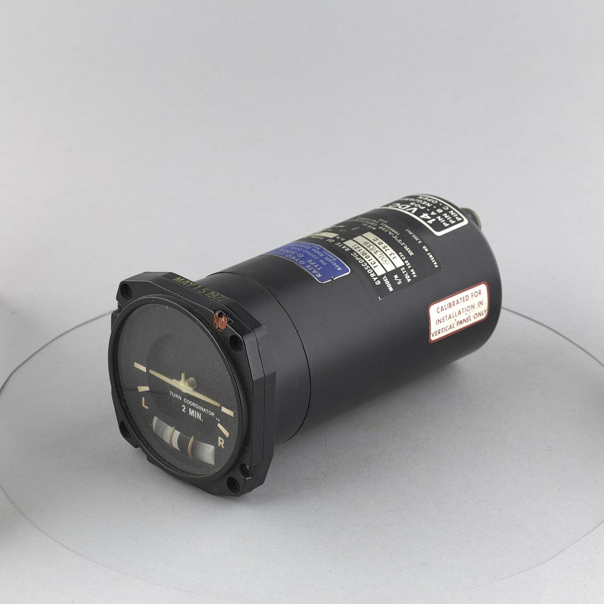 Black cylindrical instrument seen at an angle, its round face showing a small aircraft symbol with marks at each side and the words 2 MIN.