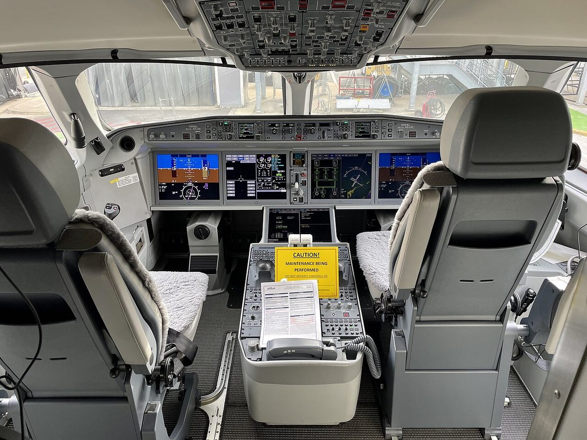 An airliner flight deck seen from behind the two seats: four large display screens across the main panel, the centre pedestal between the seats and the overhead panel above.