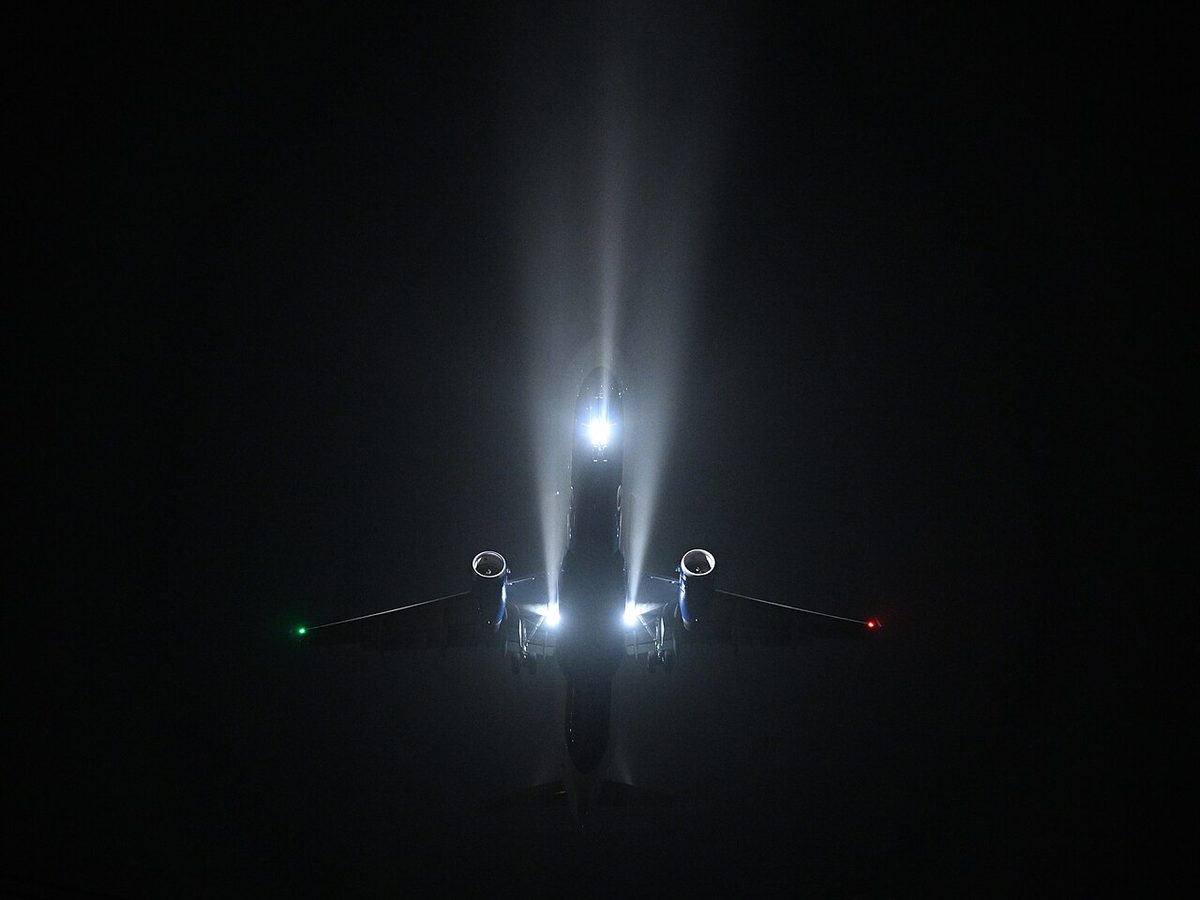An airliner seen from below at night in fog, with bright beams of light shining forward from beneath its wing roots and from its nose, and a red and a green light at the wingtips.