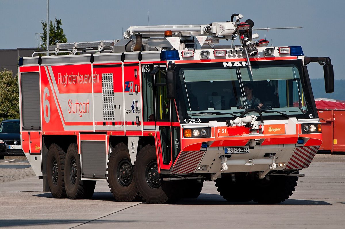 A red and white four-axle airport fire truck with a water cannon on its roof and blue lamps above the cab, on an apron.
