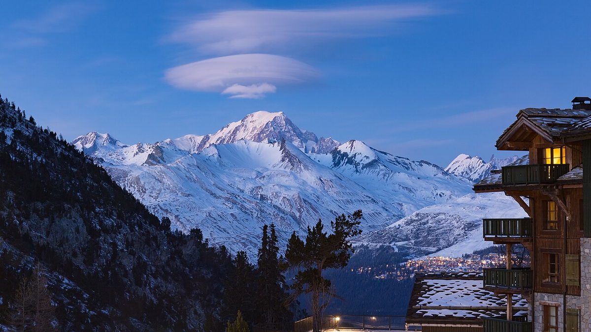 Two smooth lens-shaped clouds, one above the other, in a twilight sky over snow-covered mountains, with a lit chalet on the right.