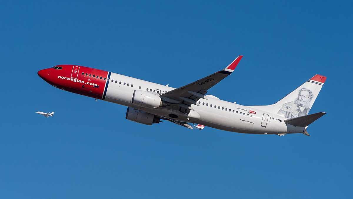A white Boeing 737 with a red nose and a portrait on its tail climbing away against a deep blue sky, landing gear up.