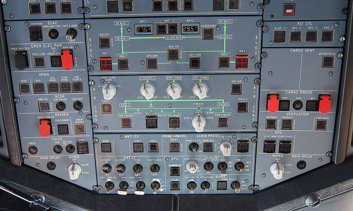 The overhead panel of an airliner cockpit, with rows of gray system panels; along the bottom edge a row of toggle switches and rotary knobs.