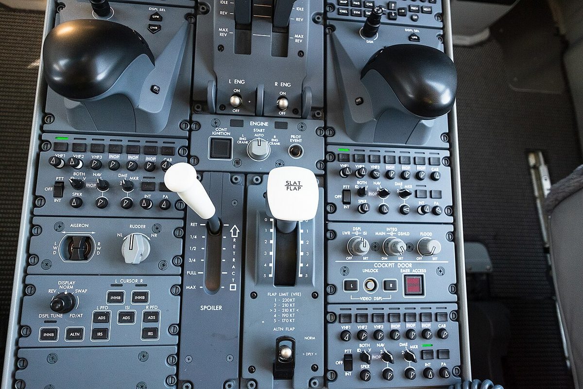 Close-up of an airliner's centre pedestal from above: thrust levers at the top, a white spoiler lever and a white slat/flap lever with a speed placard below it.