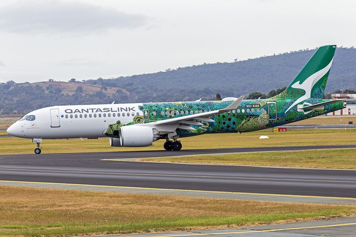 An Airbus A220-300 with QANTASLINK titles and a green patterned livery on its rear fuselage and tail, registered VH-X4A, taxiing at Canberra Airport, its nose gear and main gear on the ground.