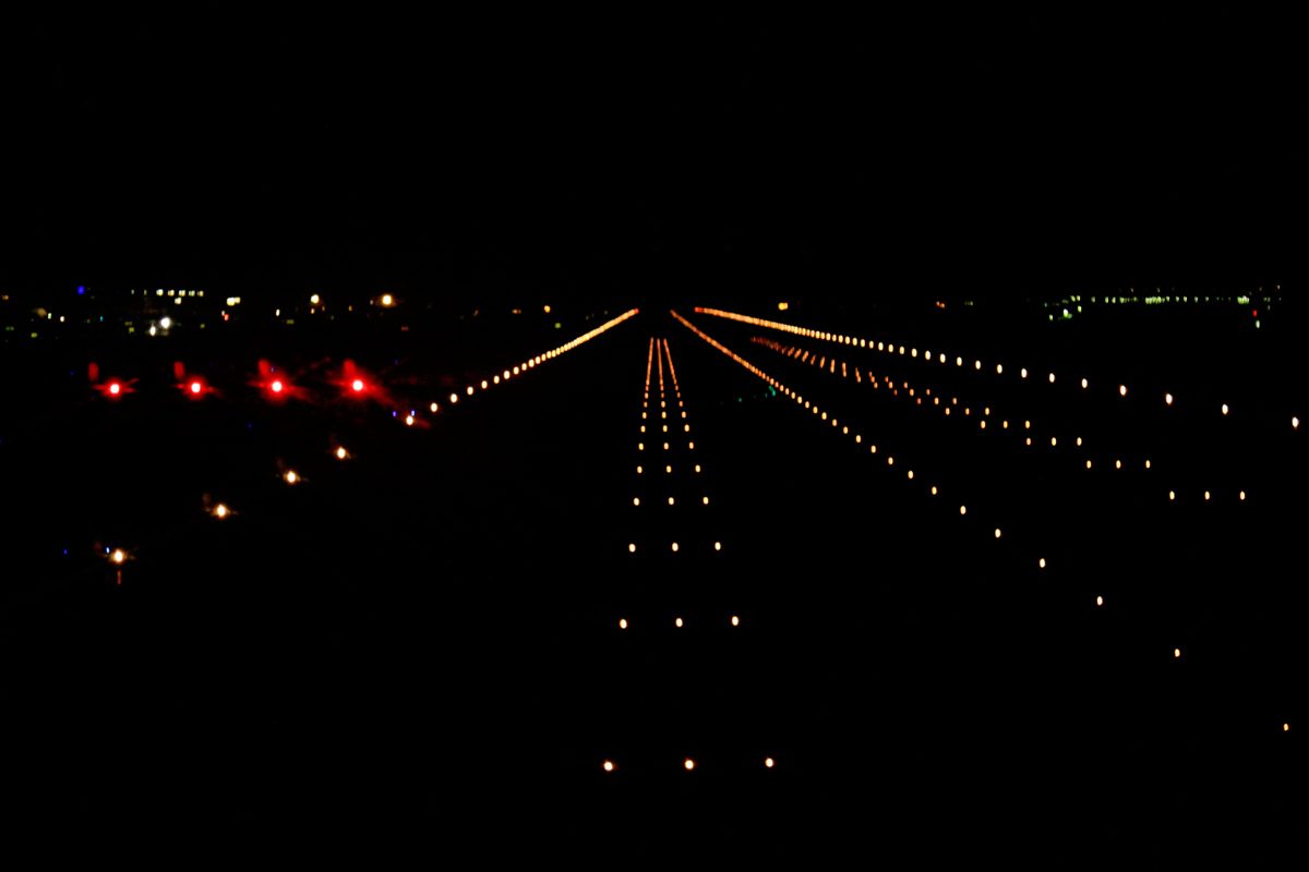 Night view along a runway from its approach end, with short bars of lights on the centre line in the foreground, lines of edge lights converging to the horizon and four red lights to the left.