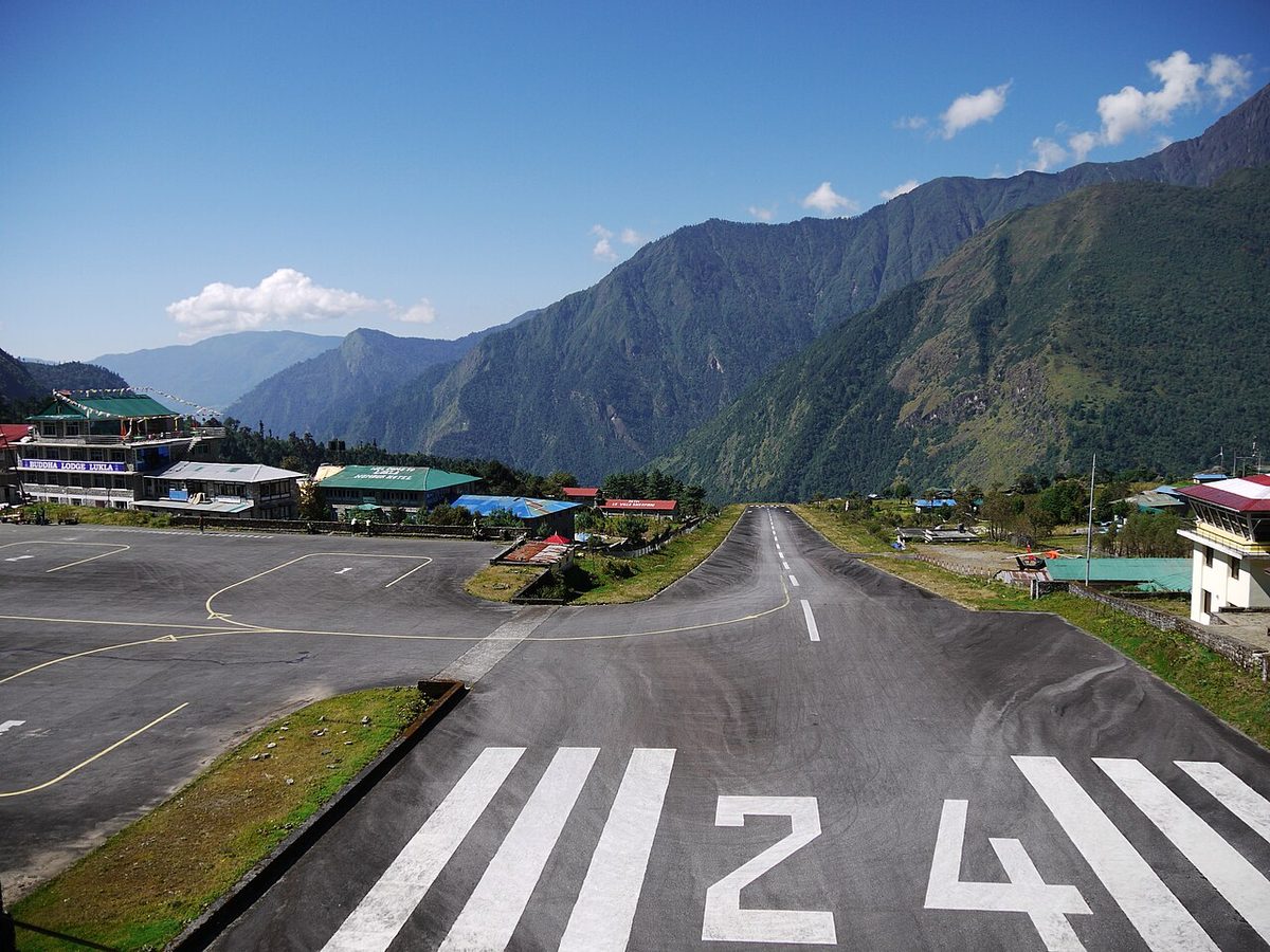 View along a runway from its end, the number 24 in the foreground and the runway dropping away down a hillside toward a mountain valley.