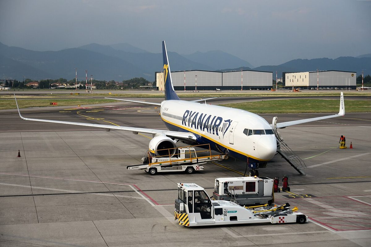 A Ryanair Boeing 737 on an apron stand with boarding stairs at the front door and a belt loader at the forward hold; in the foreground a ground power unit, its black cable running up to the lower fuselage beside the nose gear, and a tug.