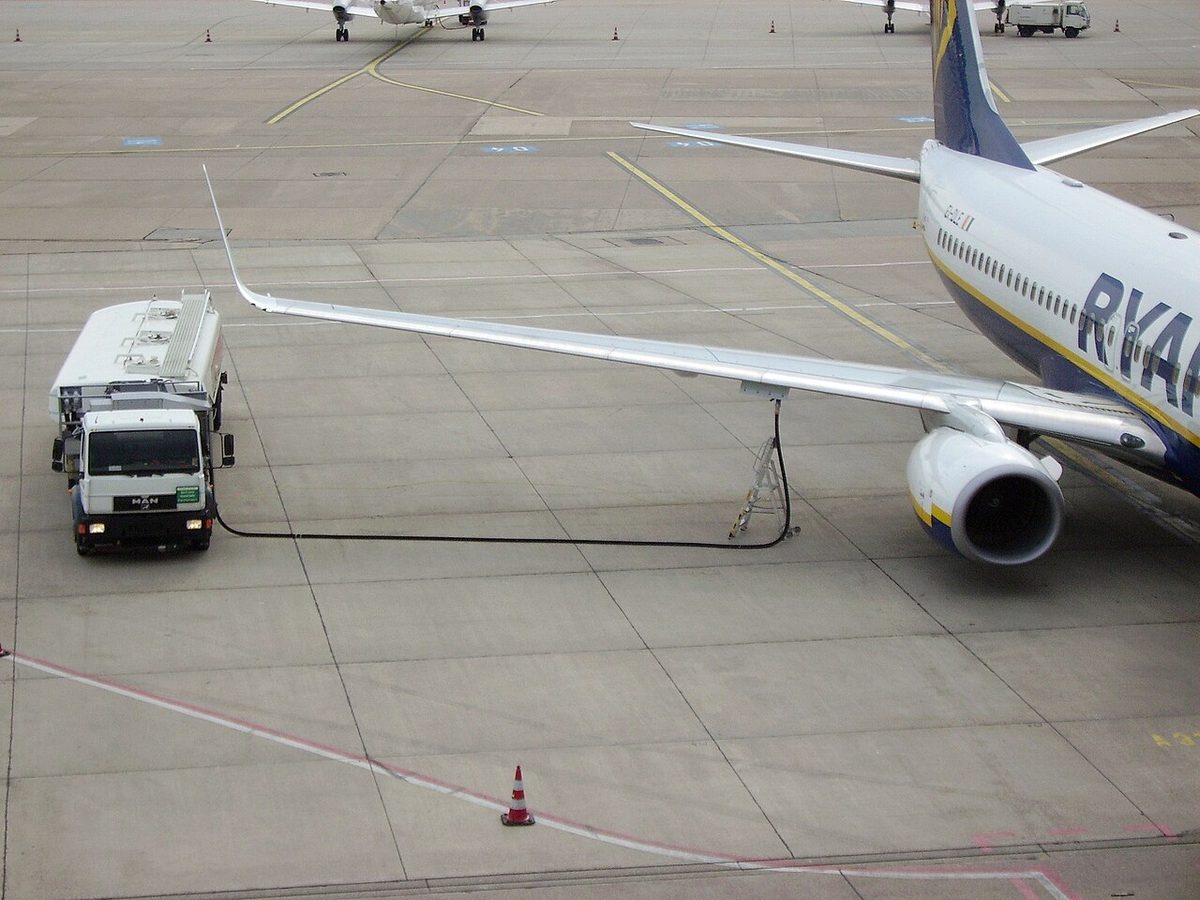 A Boeing 737 on an apron seen from above, a fuel tanker truck beside it with its hose running up to the underside of the wing just outboard of the engine.