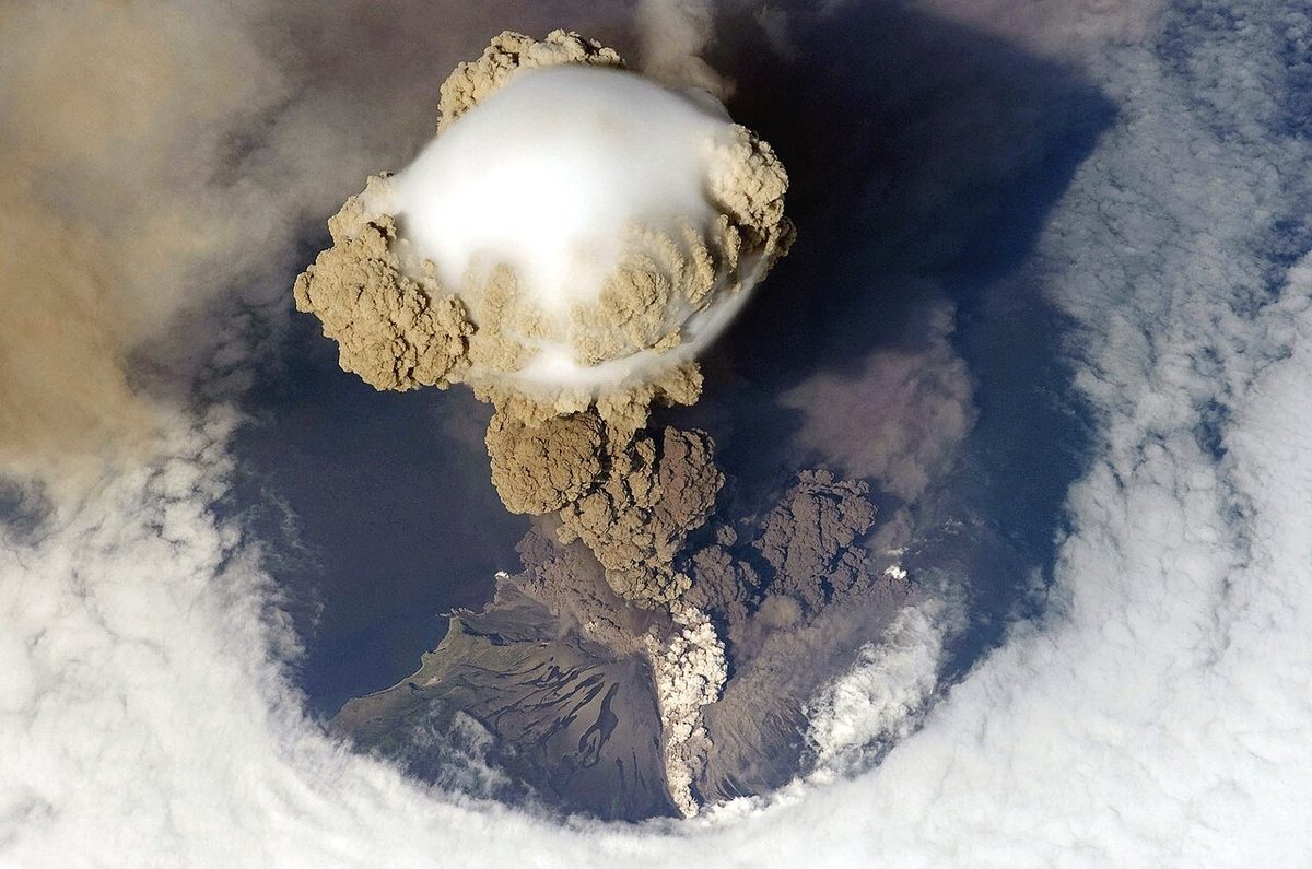 Seen from high above, a brown column of volcanic ash rising from an island through a ring of white cloud, topped by a smooth white cap.