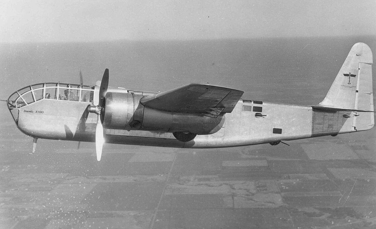 Black-and-white photo of a twin-engined aircraft with a glazed nose flying over farmland, the propeller on the nearer engine stopped.