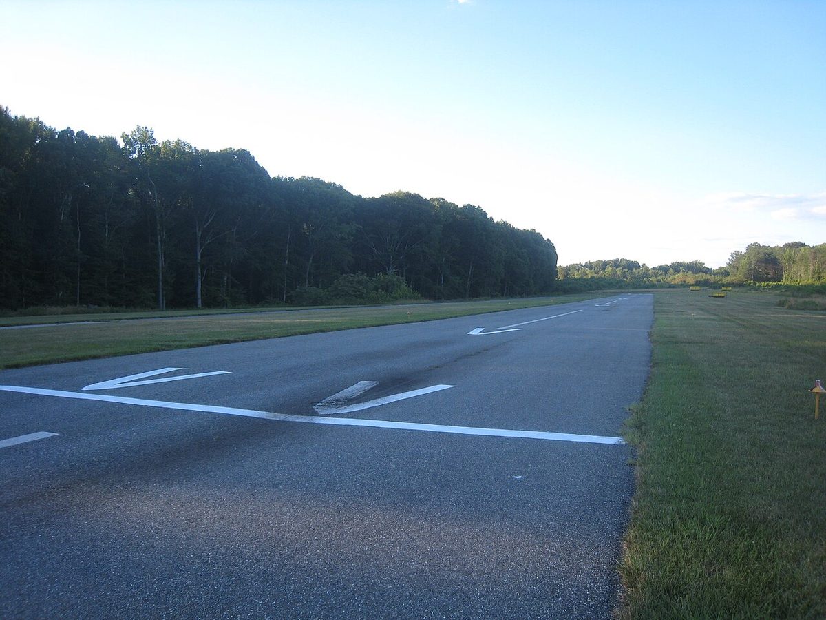 A narrow paved runway seen from ground level, with a white line painted across it and white arrowheads on the surface just before the line.