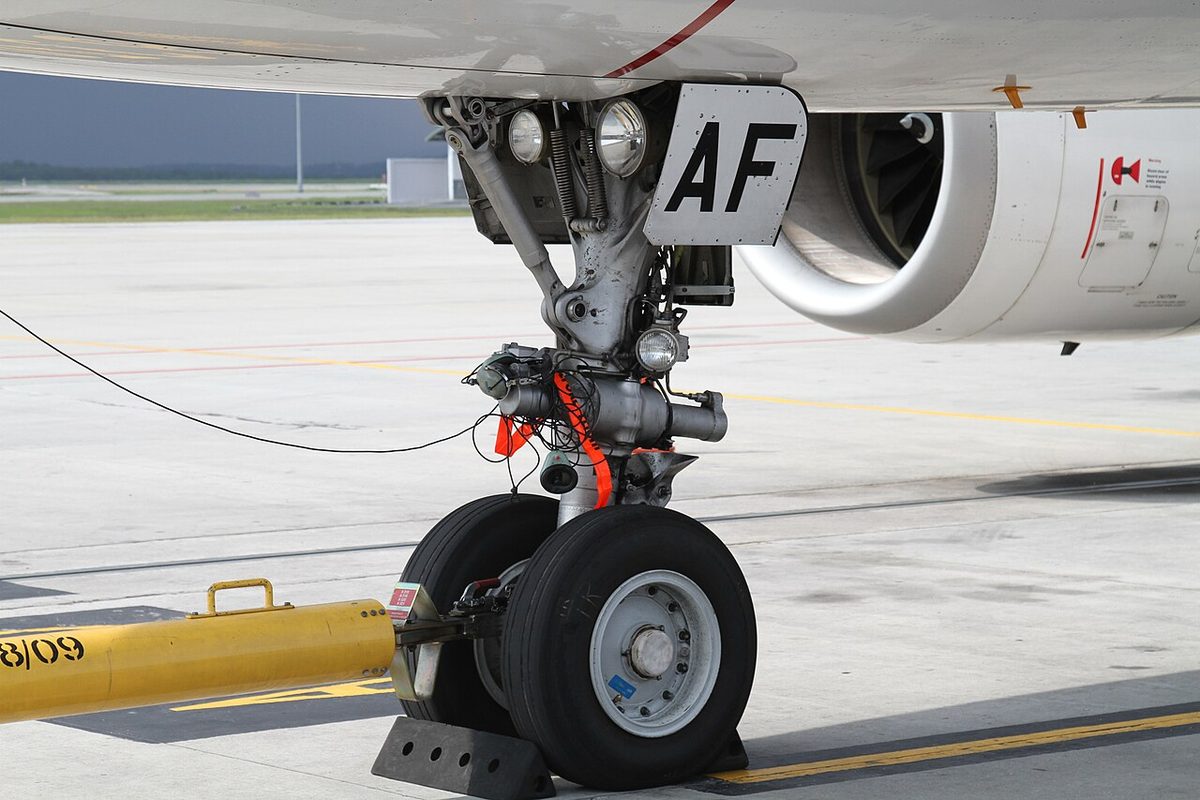 Close view of an airliner nose gear on the apron: twin wheels, a red-flagged pin on the leg, a yellow tow bar and two lamps near the top.