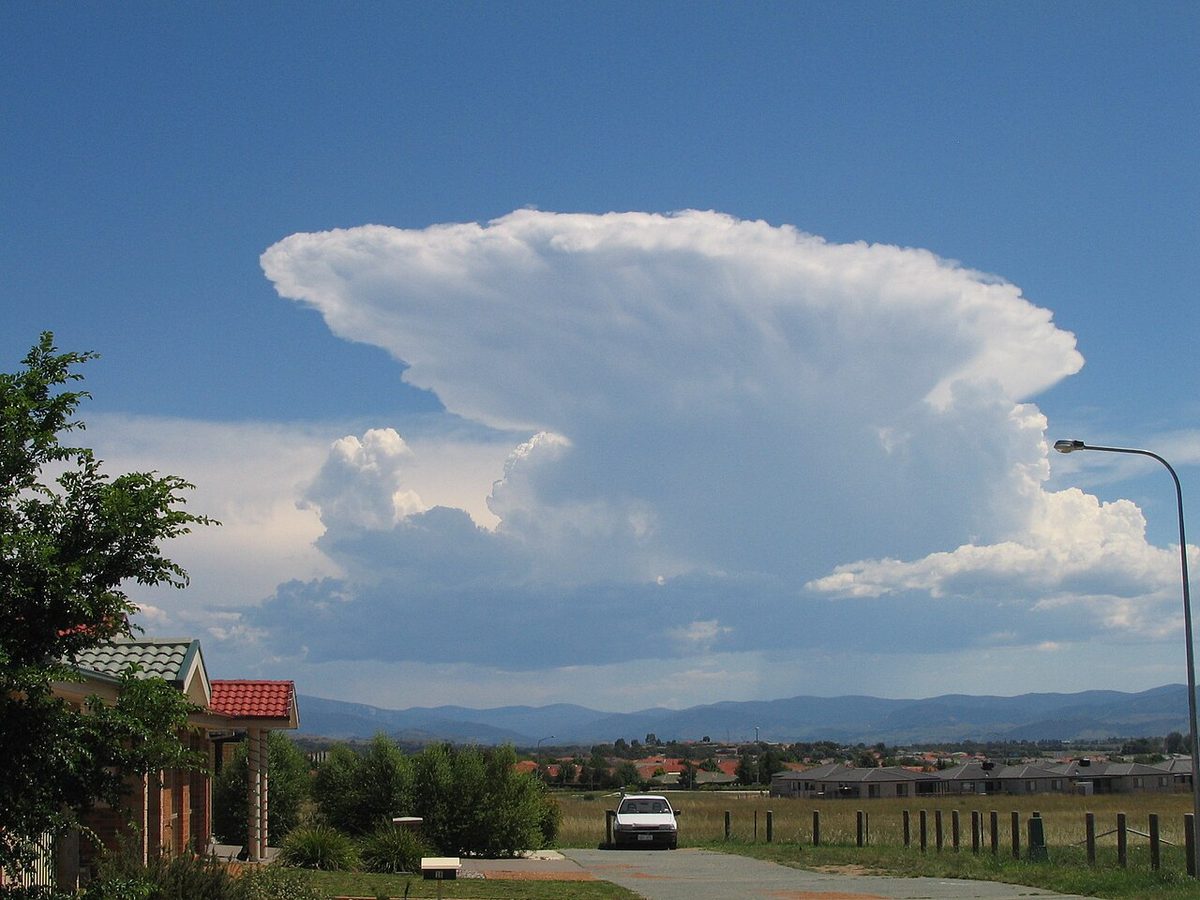 A towering white storm cloud with a flat anvil top spreading out over clear sky, above a rural road, a fence and distant hills.