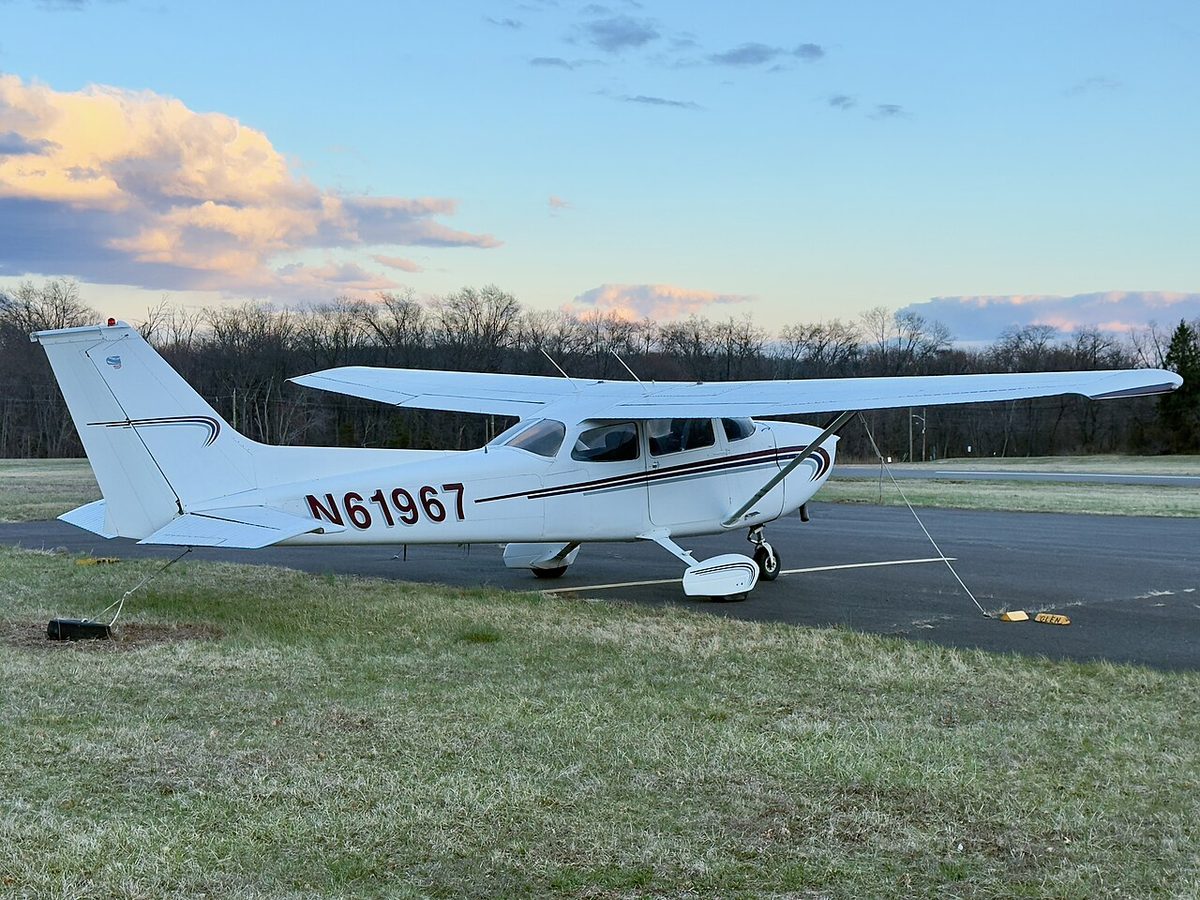 A white high-wing Cessna 172 registered N61967 parked on grass beside a paved apron under a blue evening sky.