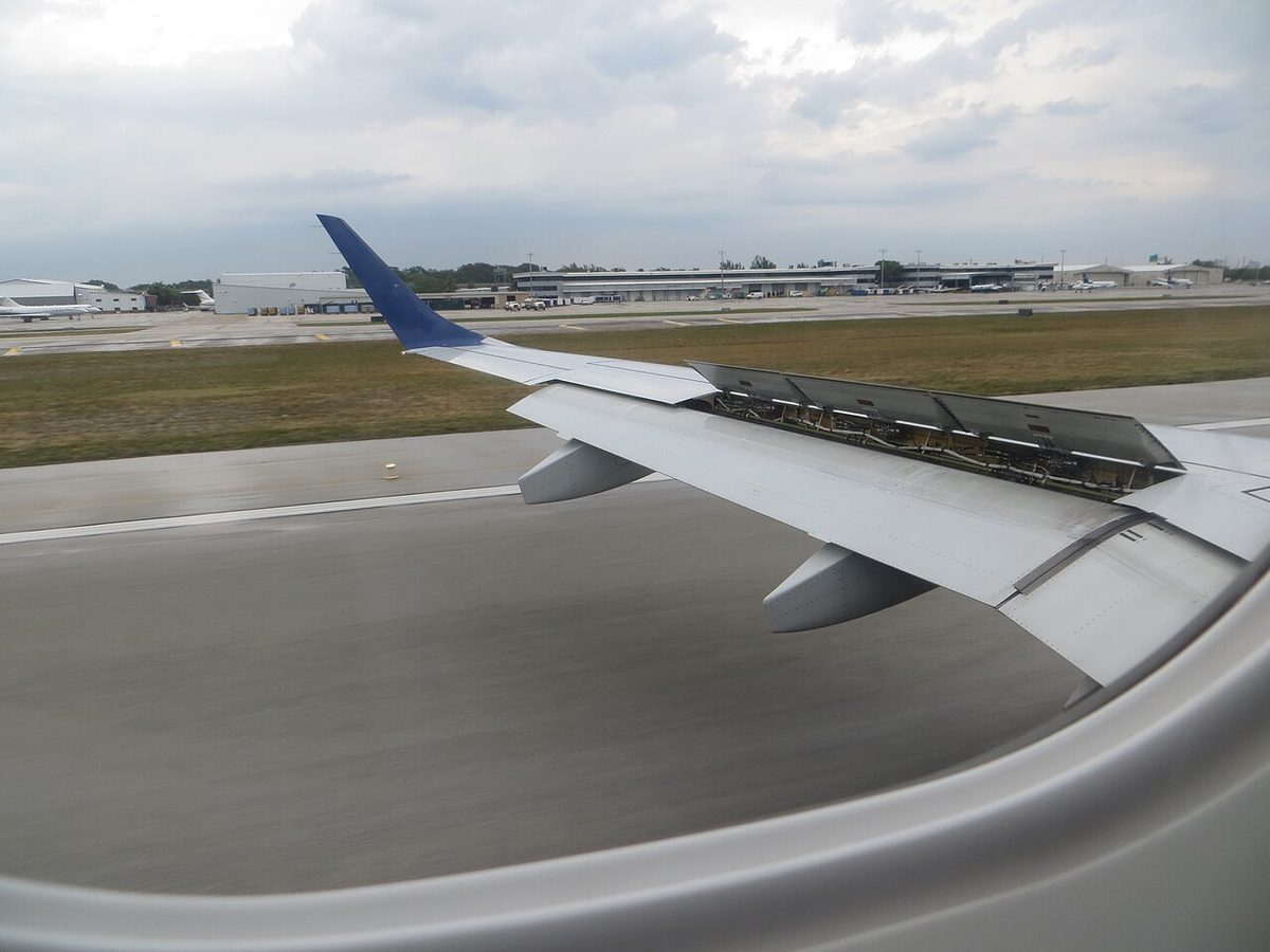 View through a cabin window of a jet's wing on the runway after landing, with a row of spoiler panels raised along the upper surface.