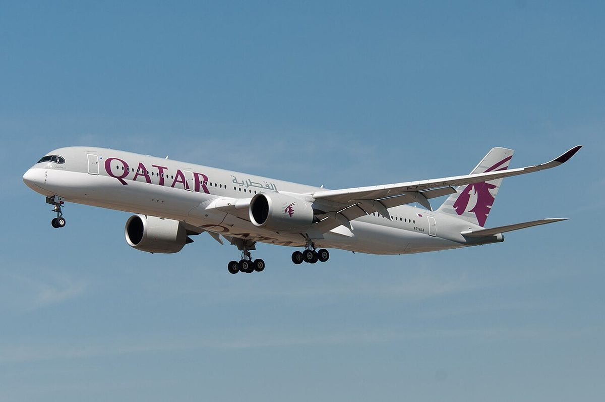A grey and white Qatar Airways Airbus A350 with a maroon tail on approach, landing gear down, against a blue sky.