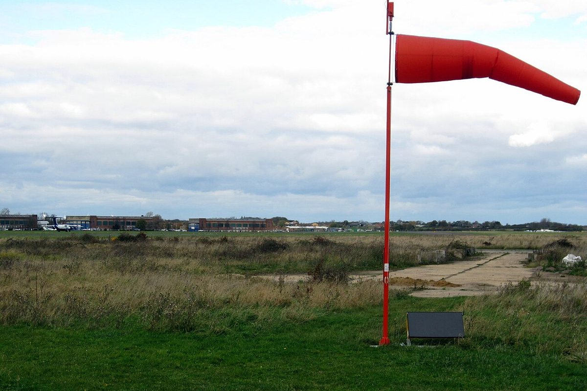 A red windsock standing out almost horizontally from its pole on a grass airfield.