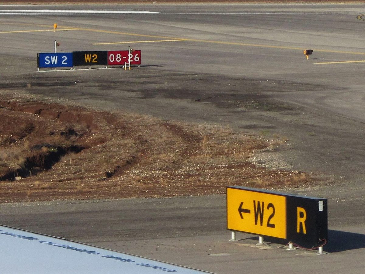 A yellow sign with a left arrow and W2 next to a black sign with a yellow R beside a taxiway, with more signs, including a red 08-26, behind.