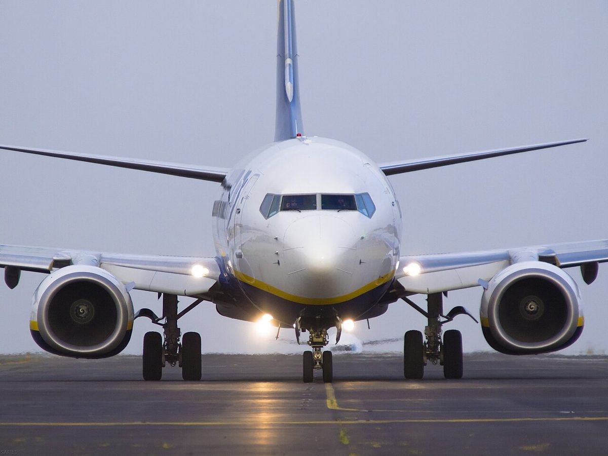 An airliner facing the camera on a taxiway in grey light, with bright lamps lit at both wing roots and under the nose.