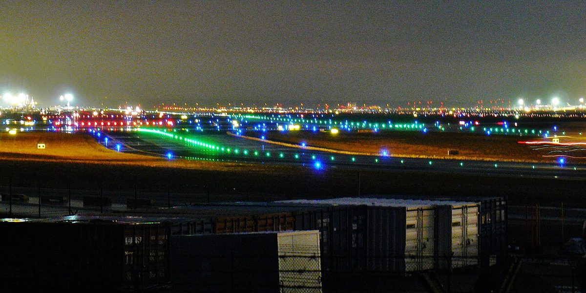 An airport at night seen over dark containers, with curving lines of green lights and scattered blue lights on the taxiways and city lights beyond.