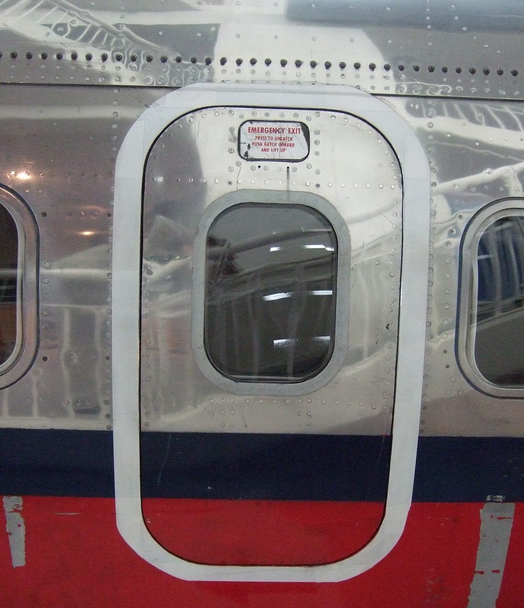 Outside of an aircraft fuselage showing a rounded rectangular hatch outline around a cabin window, with a red-lettered placard above the window.