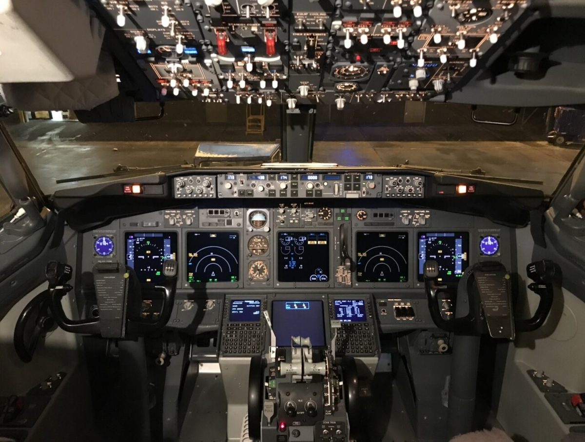 A Boeing 737-800 flight deck seen from behind the seats, with display screens across the instrument panel and the control stand between the pilots.