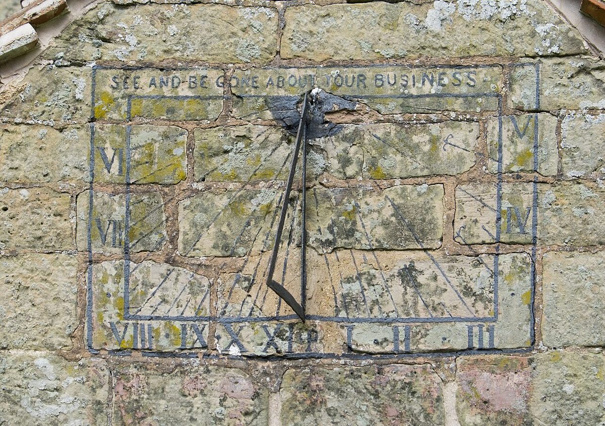 A sundial painted on the stone wall of a church porch: hour lines radiating from a metal pointer, Roman hour numerals round the edge and the motto See and be gone about your business.