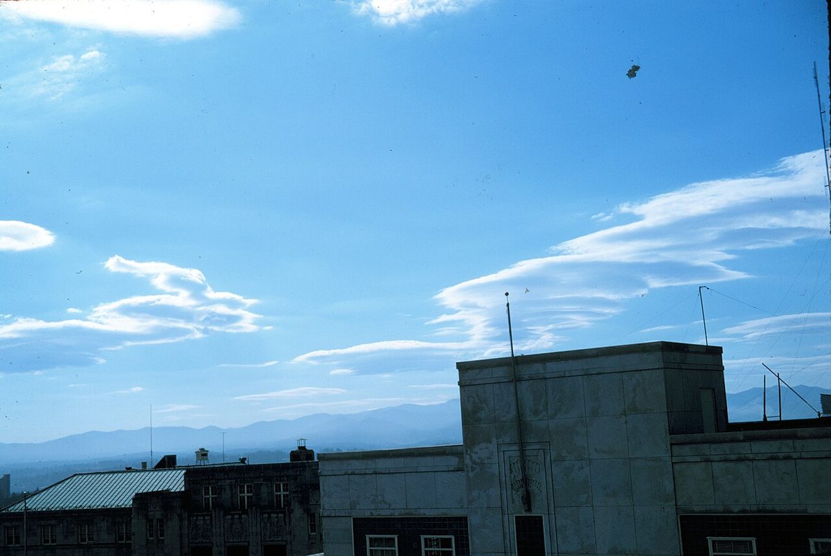 Smooth, lens-shaped clouds above a distant mountain range, seen over rooftops.
